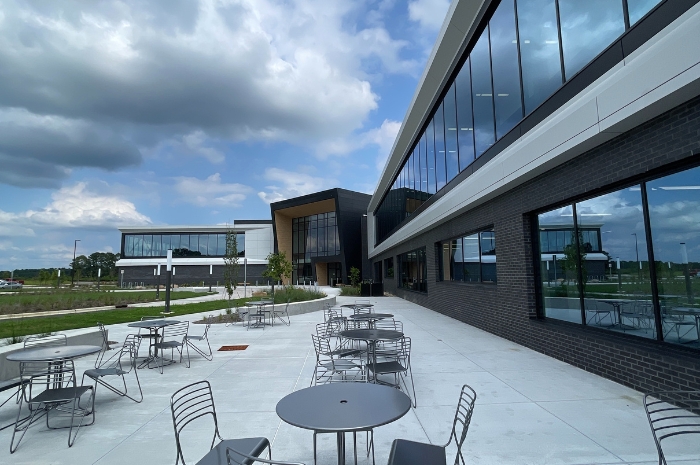 A modern campus building with large glass windows and an outdoor patio area furnished with metal tables and chairs under a partly cloudy sky.