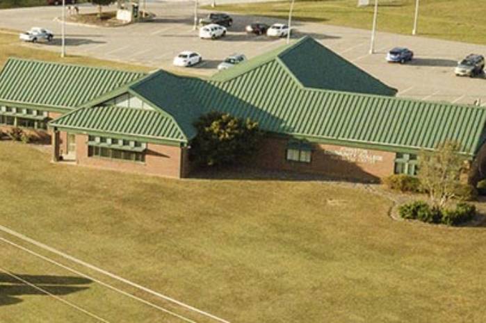 An aerial view of a one story brick campus building with green metal roofing, surrounded by open grass and a parking lot with several cars.