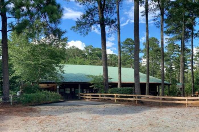 A lodge-style building with a green metal roof sits among tall pine trees, with a gravel path and wooden fence leading to the entrance of the Educational Center on a sunny day.