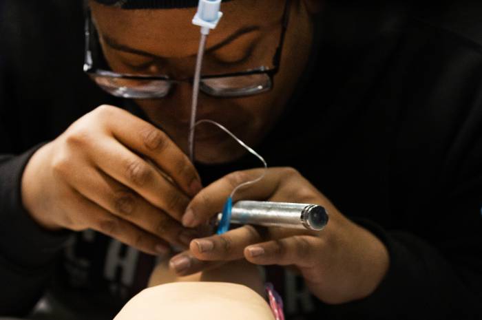 A student practices a medical airway procedure on an infant manikin, carefully inserting a tube while leaning close to the model.