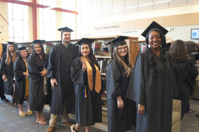 A group of students in black graduation caps and gowns stand in a library, smiling and posing in a line before their ceremony.